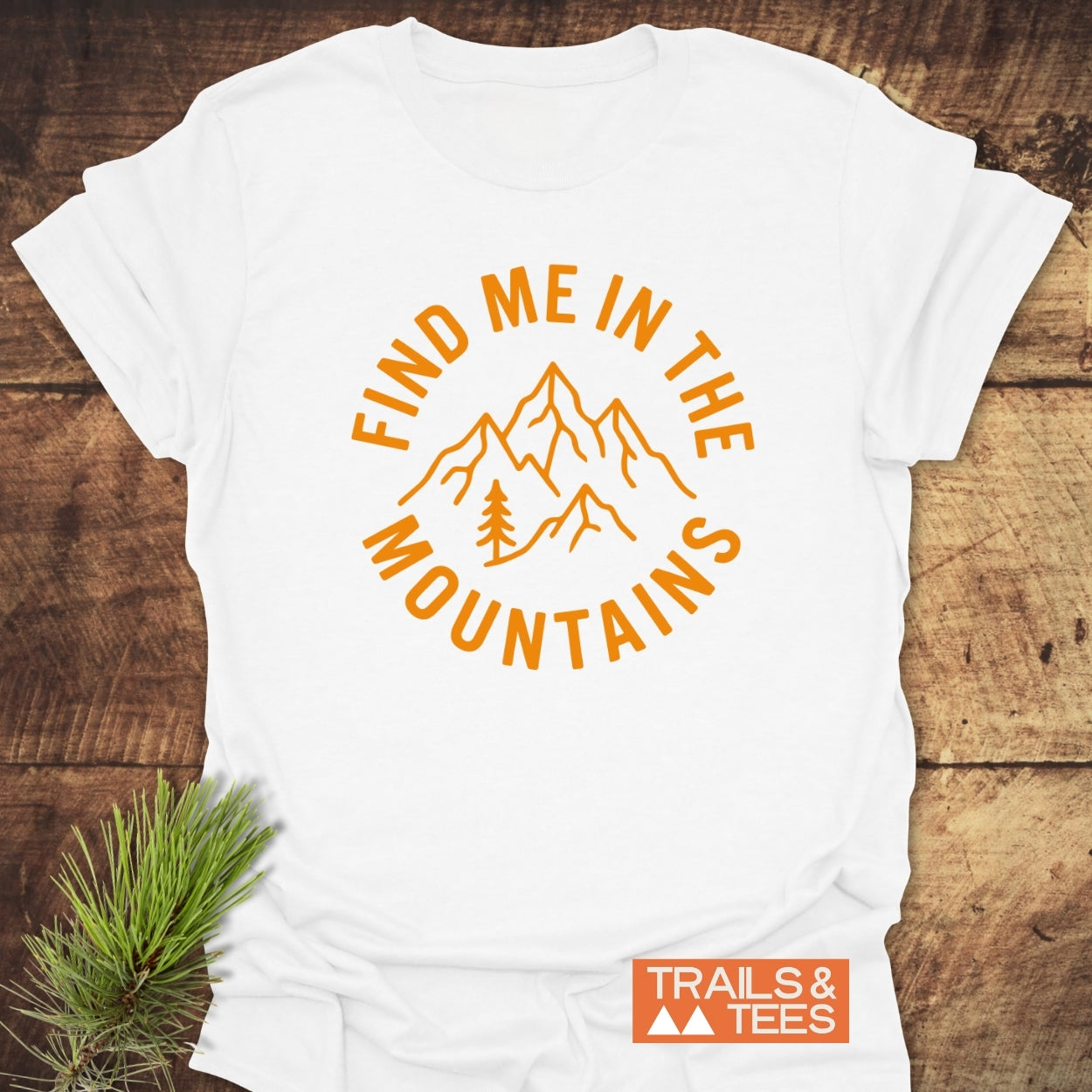 Find Me In The Mountains T-Shirt featuring an orange mountain and pine tree design, surrounded by text, on a flat-lay wood surface with a pine sprig.