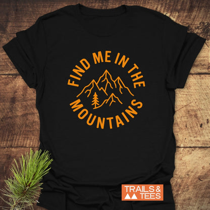 Find Me In The Mountains T-Shirt featuring bold orange mountain peaks and pine tree line-art on a black T-shirt, with “TRAILS & TEES” logo, showcasing outdoor adventure spirit on a wooden surface.