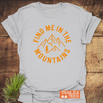 Find Me In The Mountains T-Shirt featuring bold orange mountain peaks and pine tree design, styled with a pine sprig. Laid on a wooden surface, includes TRAILS & TEES logo.