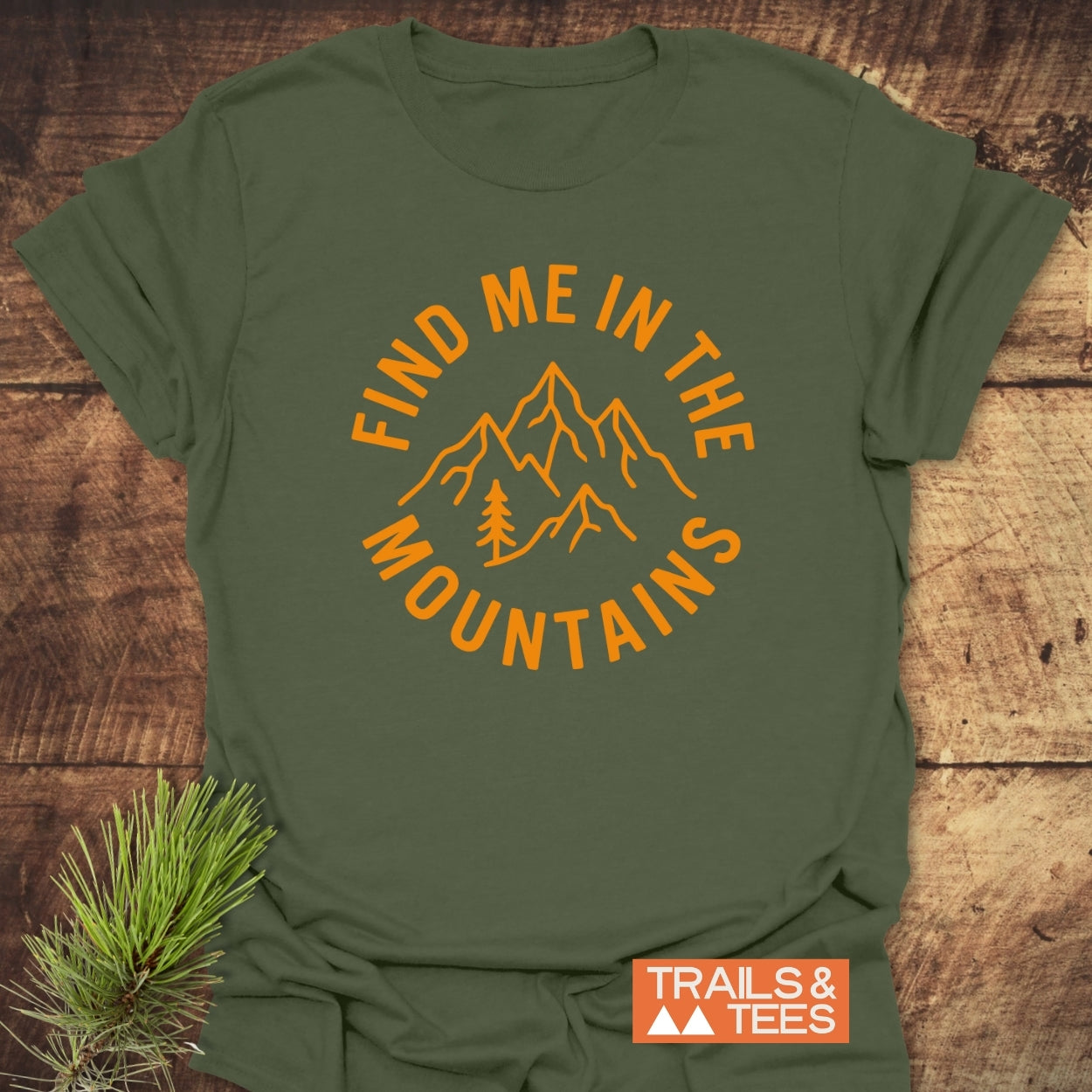 Find Me In The Mountains T-Shirt on a wooden surface, featuring an orange graphic of mountains and a pine tree. Includes a “TRAILS & TEES” logo tag and a pine branch prop for outdoor appeal.