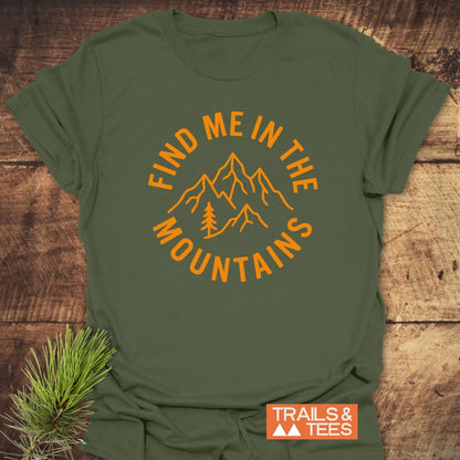 Find Me In The Mountains T-Shirt on a wooden surface, featuring an orange graphic of mountains and a pine tree. Includes a “TRAILS & TEES” logo tag and a pine branch prop for outdoor appeal.