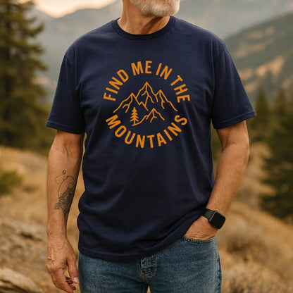 Man wearing Find Me In The Mountains T-Shirt with orange mountain and pine tree design, showcasing outdoor adventure spirit. Accessorized with a smartwatch and a forearm tattoo, set in a mountain backdrop.