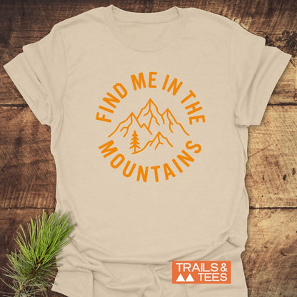 Find Me In The Mountains T-Shirt: Light-heather-tan crew-neck with orange mountain and pine design, laid on rustic wood. Perfect for outdoor enthusiasts, offering comfort and durability with eco-friendly, on-demand printing.