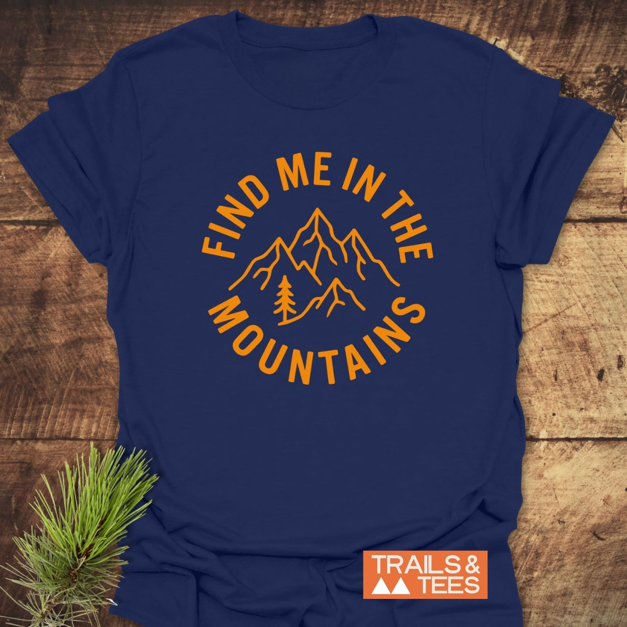 Find Me In The Mountains T-Shirt: Navy-blue tee with bold orange mountain design and FIND ME IN THE MOUNTAINS text. Features durable prints, Trails & Tees logo, and pine sprig accent.