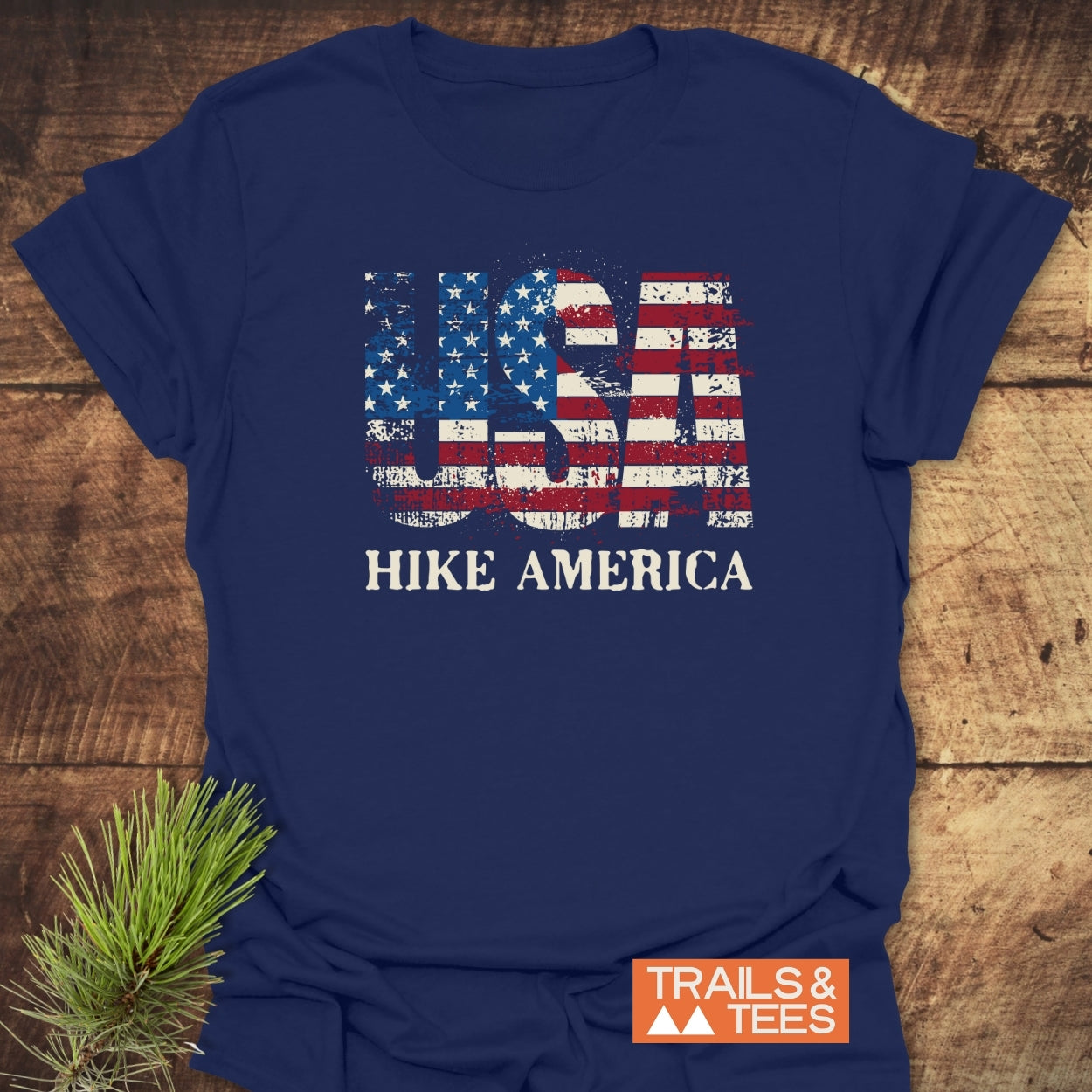 Navy-blue Hike America T-Shirt with distressed USA stars-and-stripes print, HIKE AMERICA text below, and TRAILS & TEES logo, laid flat on wood with a pine sprig beside it.