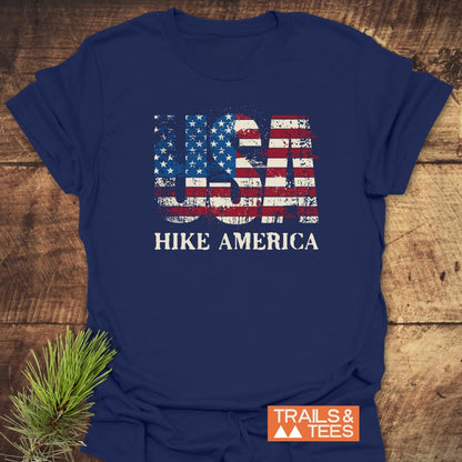 Navy-blue Hike America T-Shirt with distressed USA stars-and-stripes print, HIKE AMERICA text below, and TRAILS & TEES logo, laid flat on wood with a pine sprig beside it.