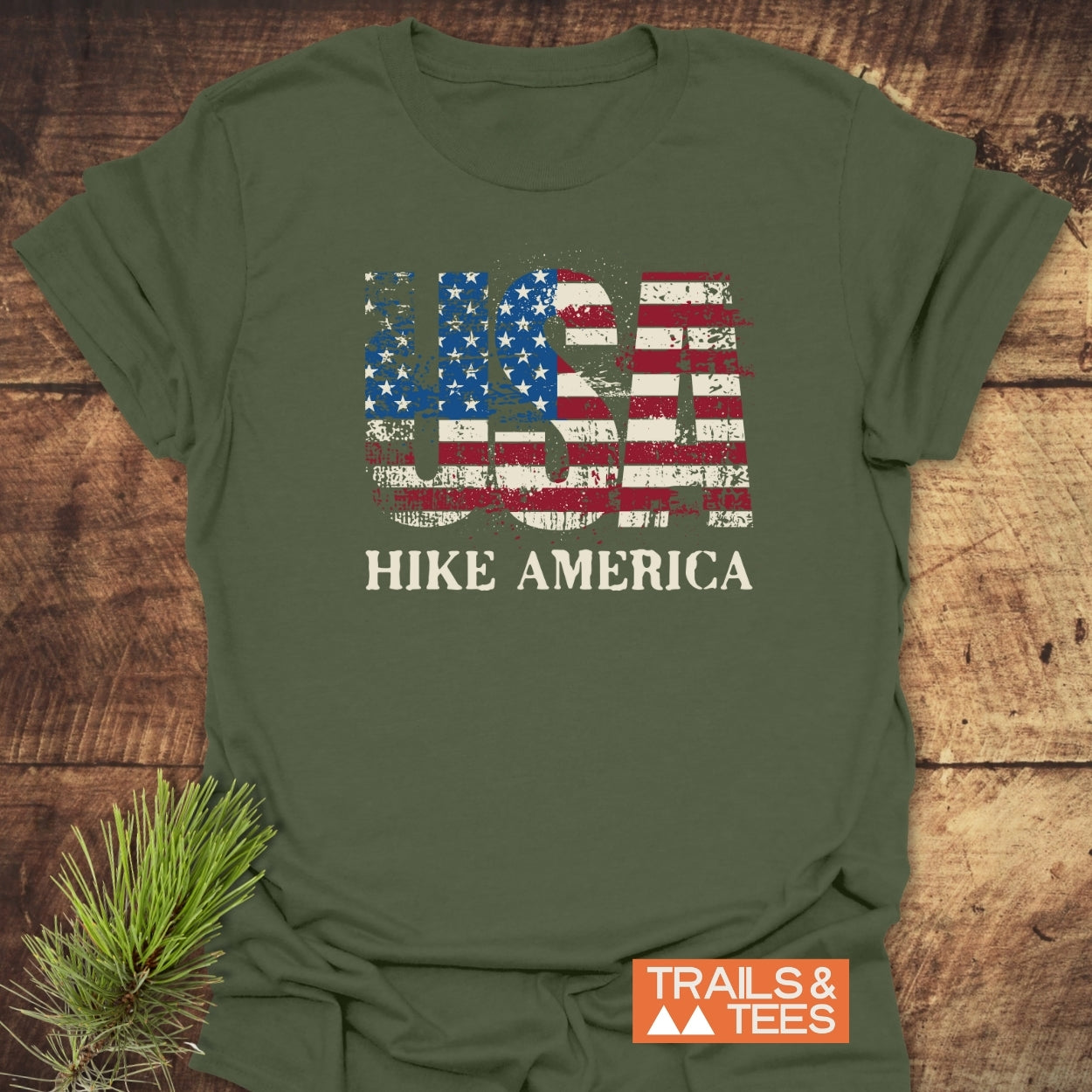Hike America T-Shirt with a distressed USA stars-and-stripes graphic and slogan. Olive-green, crafted from soft ring-spun cotton, styled with a pine branch and TRAILS & TEES logo.