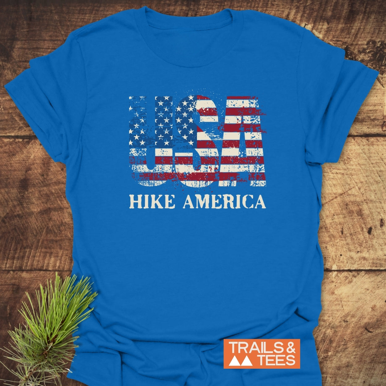 Hike America T-Shirt with distressed USA flag graphic and HIKE AMERICA text, laid flat on wood. Soft ring-spun cotton, ideal for outdoor enthusiasts, featuring TRAILS & TEES logo with mountain icons.