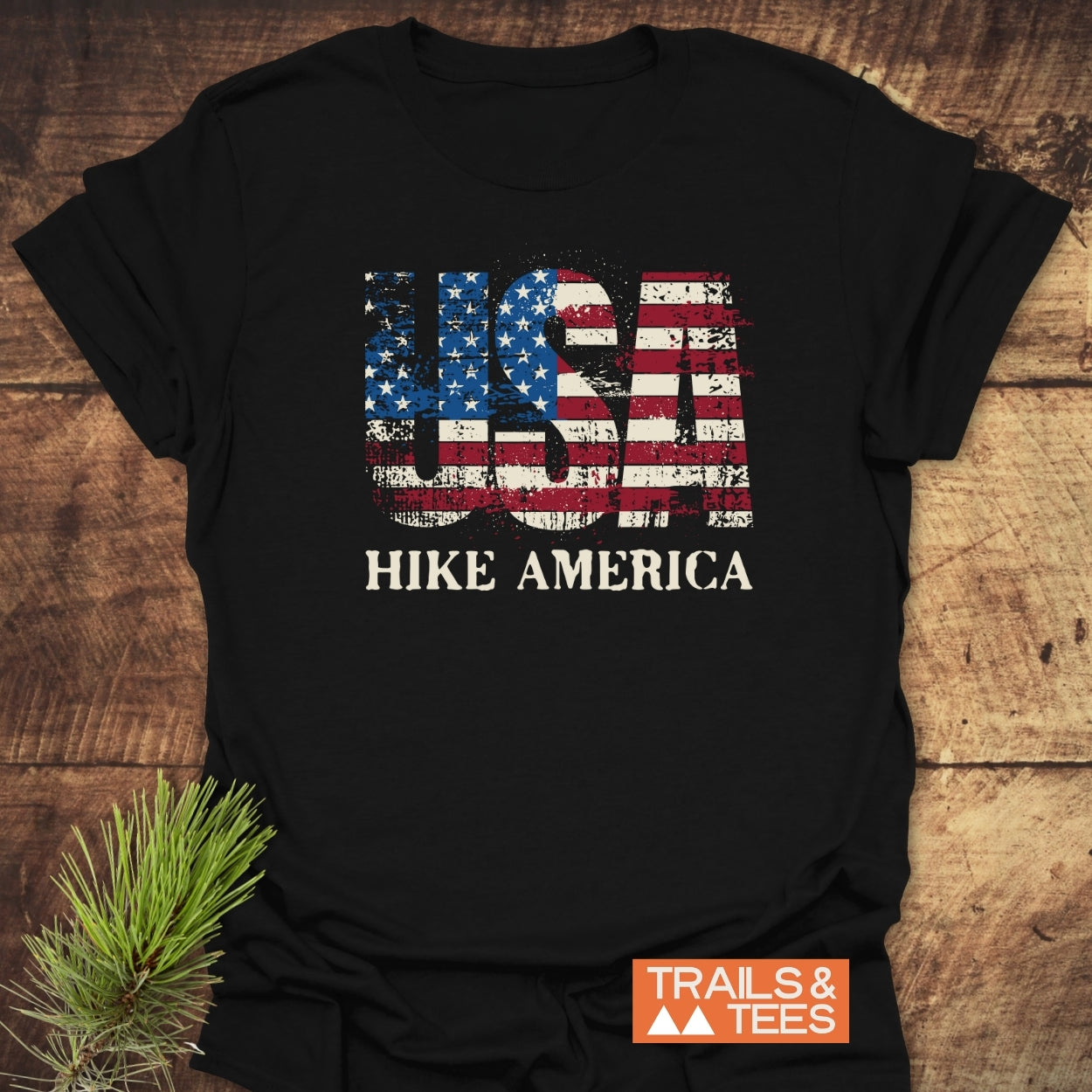 Hike America T-Shirt featuring USA stars-and-stripes print and HIKE AMERICA text, laid on wood. Includes Trails & Tees logo. Pine sprig adds rustic, outdoorsy touch.