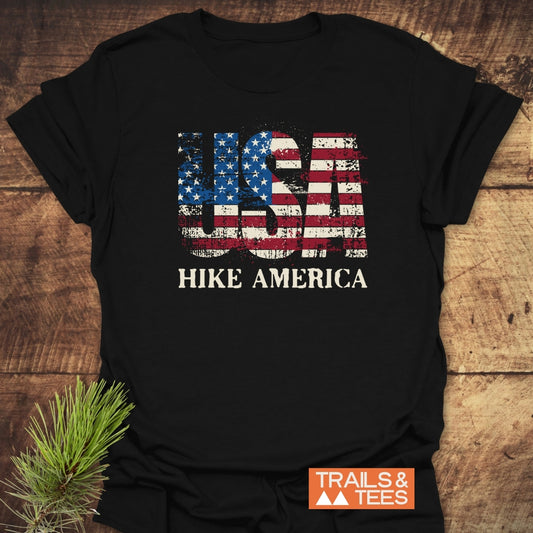 Hike America T-Shirt featuring USA stars-and-stripes print and HIKE AMERICA text, laid on wood. Includes Trails & Tees logo. Pine sprig adds rustic, outdoorsy touch.