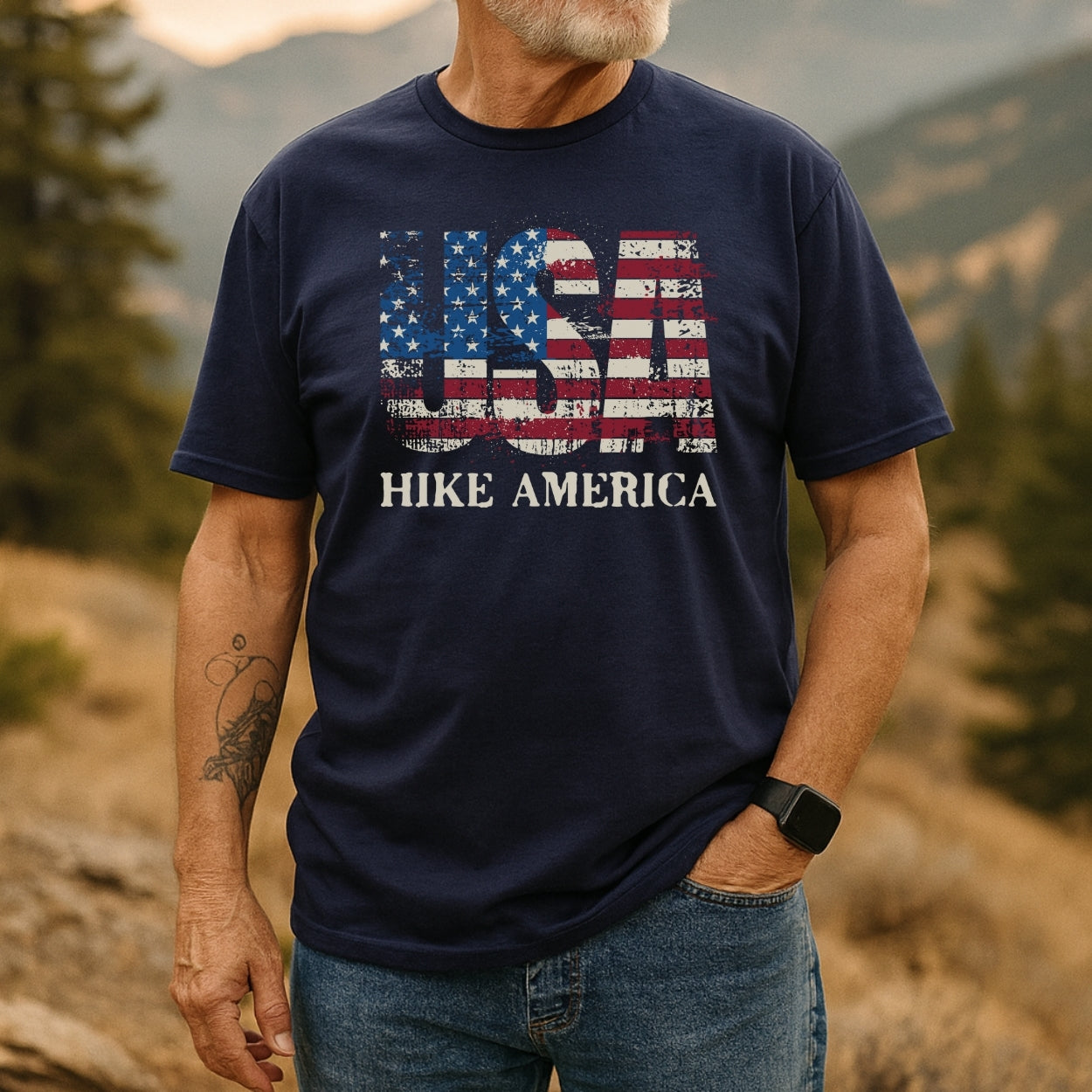 Man outdoors wearing Hike America T-Shirt with distressed American-flag motif spelling USA and HIKE AMERICA text, showcasing casual style and comfort, complemented by a smartwatch and tattoo.