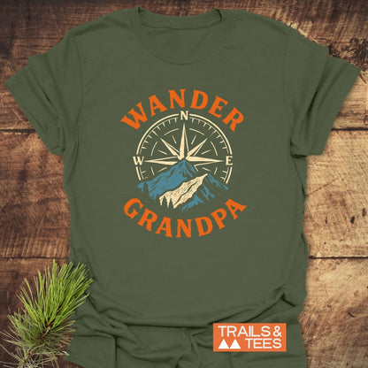 Wander Grandpa T-Shirt featuring a compass rose and mountain design, with WANDER and GRANDPA text. Displayed on a rustic surface, highlighting its adventurous theme. TRAILS & TEES logo nearby.