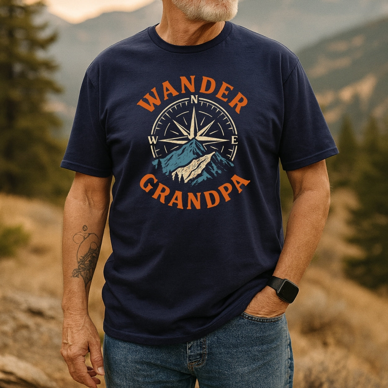 Older man outdoors in a Wander Grandpa T-Shirt, featuring a compass and mountain design. Crafted from soft cotton, ideal for adventures. Wears blue jeans, wristwatch, and has a forearm tattoo.