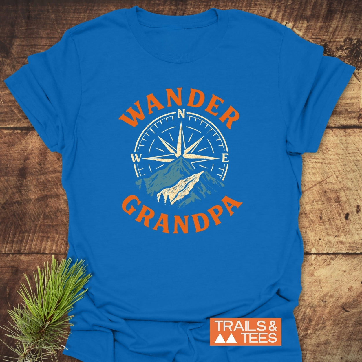 Wander Grandpa T-Shirt featuring a compass rose and mountain silhouette graphic, with WANDER and GRANDPA in bold orange. Laid on a wooden surface with a pine branch accent.