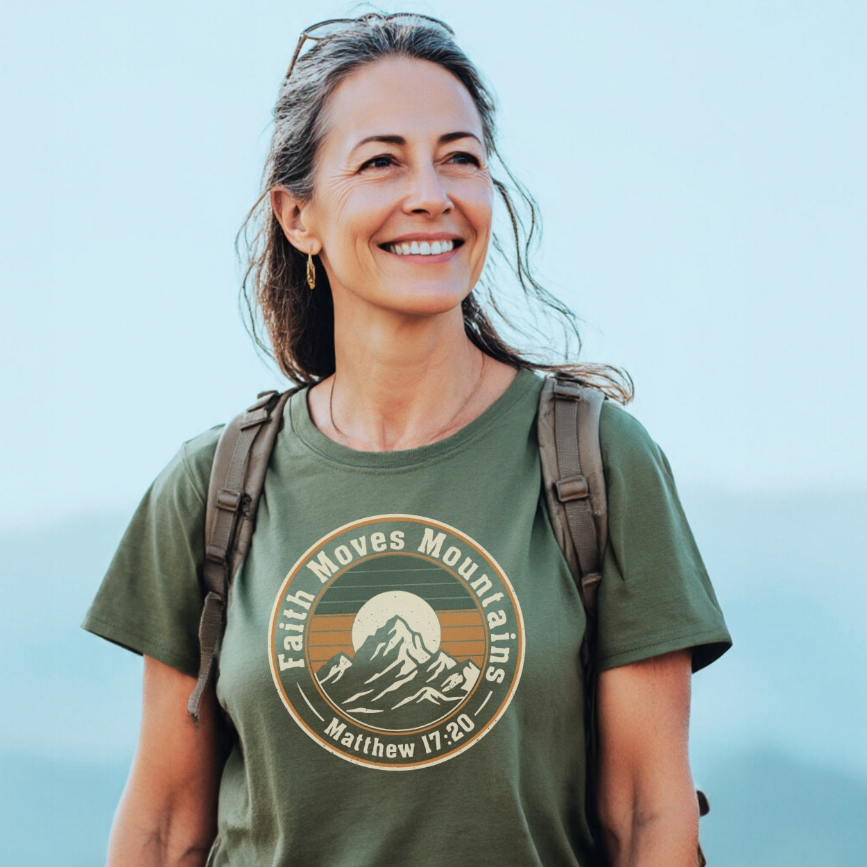 Smiling woman hiking, wearing Faith Moves Mountains T-Shirt with a mountain and sun logo, emphasizing outdoor love. Hair tied back, sunglasses, and earrings complete her trail-ready look.