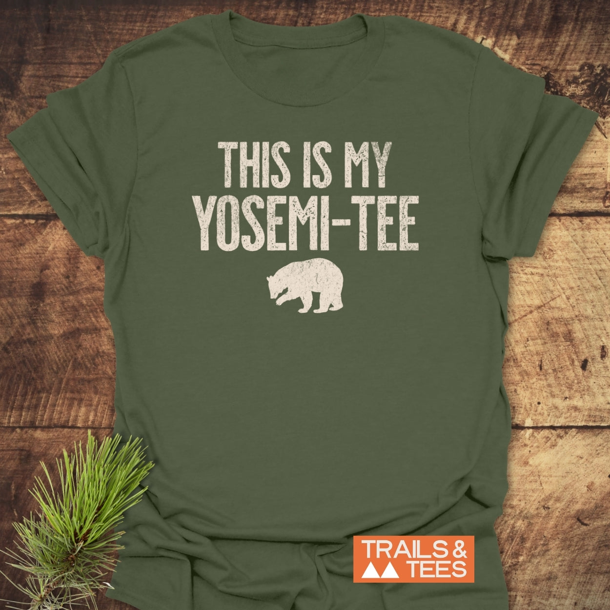 This Is My Yosemi-Tee Funny T-Shirt features a distressed white slogan and bear silhouette on olive-green fabric, styled with a pine branch beside it for an outdoorsy touch.
