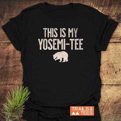 This Is My Yosemi-Tee Funny T-Shirt features a distressed white print on black fabric with a bear silhouette, lying on wood. Includes 'TRAILS & TEES' orange logo, highlighting outdoor adventure spirit.