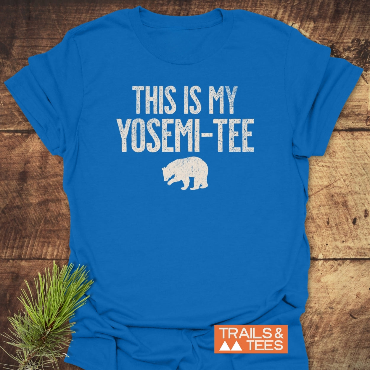 This Is My Yosemi-Tee Funny T-Shirt features white distressed lettering and a bear silhouette, showcasing outdoor-inspired design with 'TRAILS & TEES' branding, laid on a wooden surface with pine sprig.