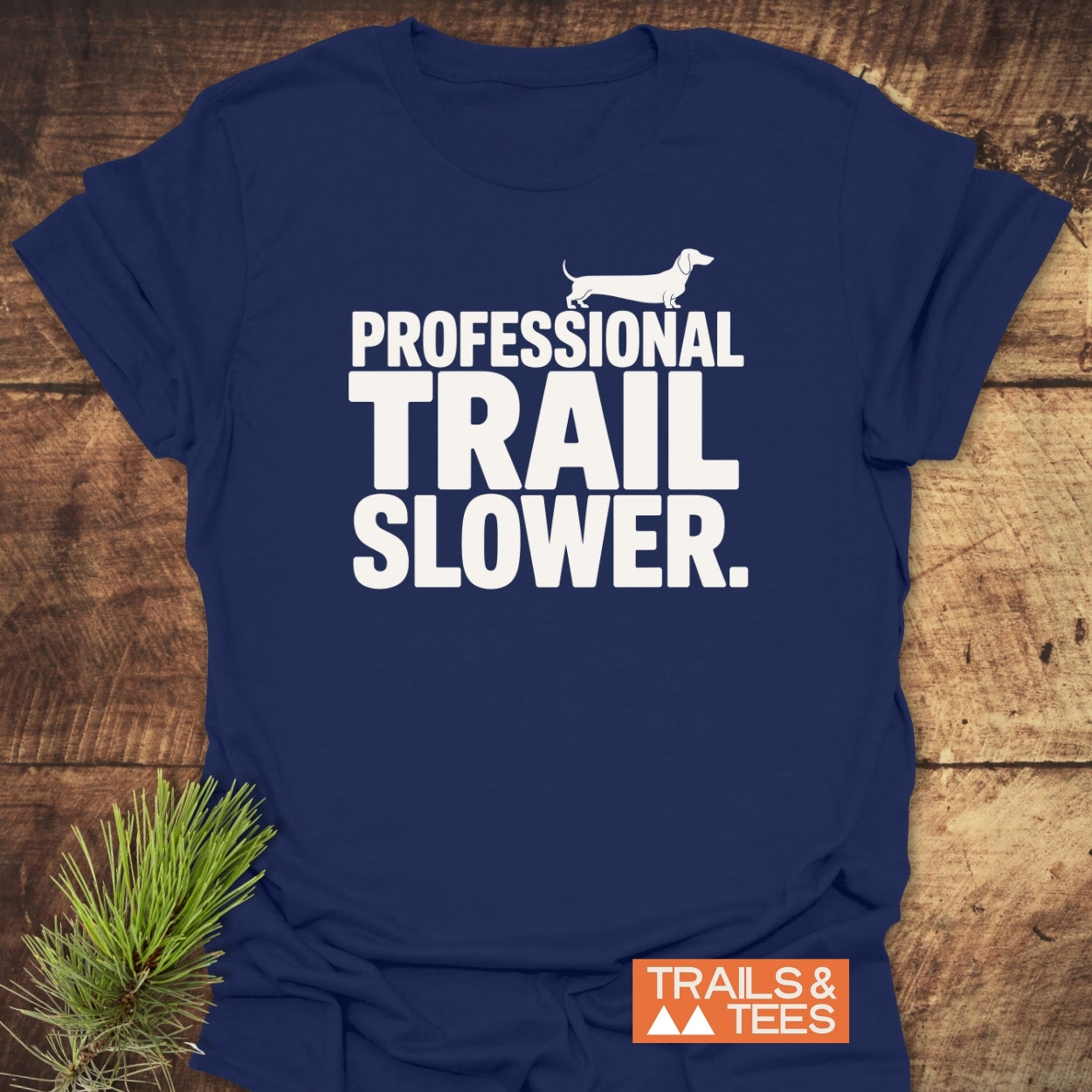 Trail Slower Dachshund T-Shirt with bold white lettering and dachshund silhouette on navy fabric, styled with pine sprigs. Features TRAILS & TEES logo, offering comfort and durability for outdoor enthusiasts.