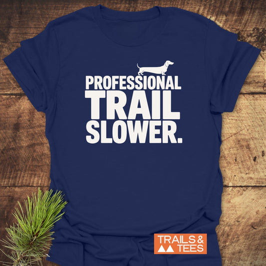 Trail Slower Dachshund T-Shirt with bold white lettering and dachshund silhouette on navy fabric, styled with pine sprigs. Features TRAILS & TEES logo, offering comfort and durability for outdoor enthusiasts.