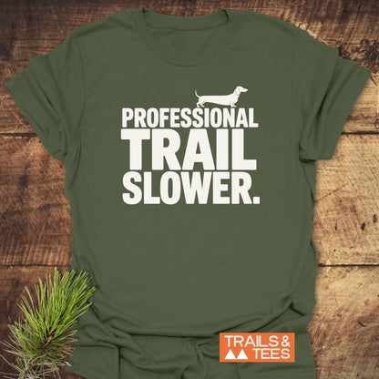 Trail Slower Dachshound T-Shirt featuring PROFESSIONAL TRAIL SLOWER in white letters with a dachshund silhouette; includes TRAILS & TEES logo. Laid on a rustic wood surface.