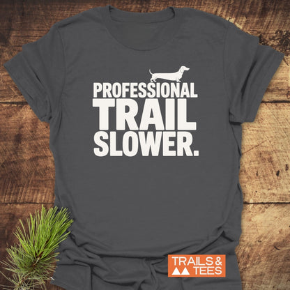 Trail Slower Dachshund T-Shirt on wooden surface with pine sprig; features white dachshund silhouette graphic and “PROFESSIONAL TRAIL SLOWER” text. Visible “TRAILS & TEES” logo in corner.