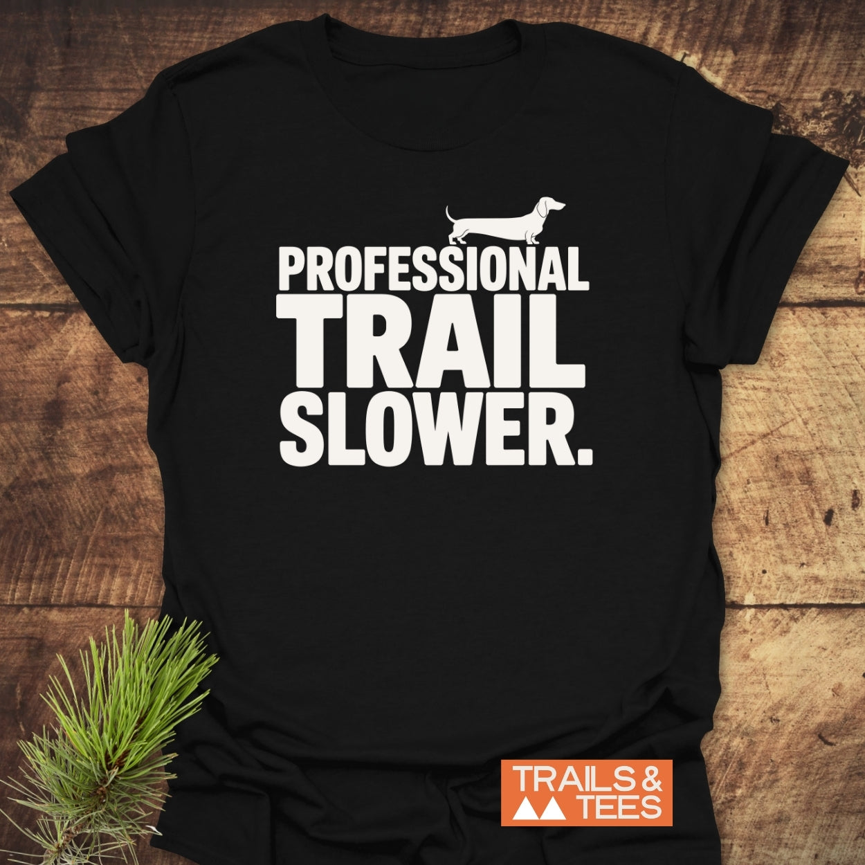Trail Slower Dachshund T-Shirt featuring bold white text and Dachshund silhouette. Displayed on wood with pine sprig, highlighting its rustic charm. Includes “TRAILS & TEES” logo in orange.