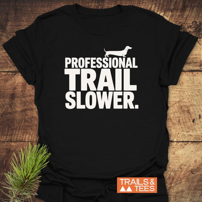 Trail Slower Dachshund T-Shirt featuring bold white text and Dachshund silhouette. Displayed on wood with pine sprig, highlighting its rustic charm. Includes “TRAILS & TEES” logo in orange.