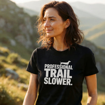 Woman wearing the Trail Slower Dachshund T-Shirt on a sunlit hillside, showcasing its dachshund silhouette and bold text. The black tee offers comfort with ring-spun cotton and durable prints.
