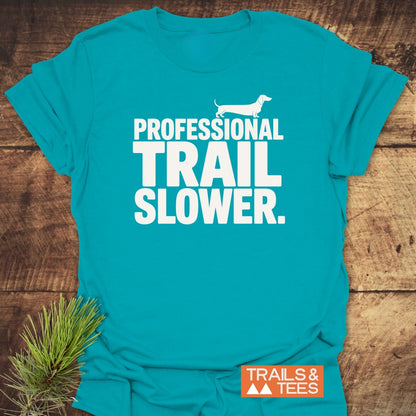 Trail Slower Dachshund T-Shirt featuring a white dachshund silhouette and bold text on teal, laid flat with a sprig of pine. Includes TRAILS & TEES logo in the bottom corner.