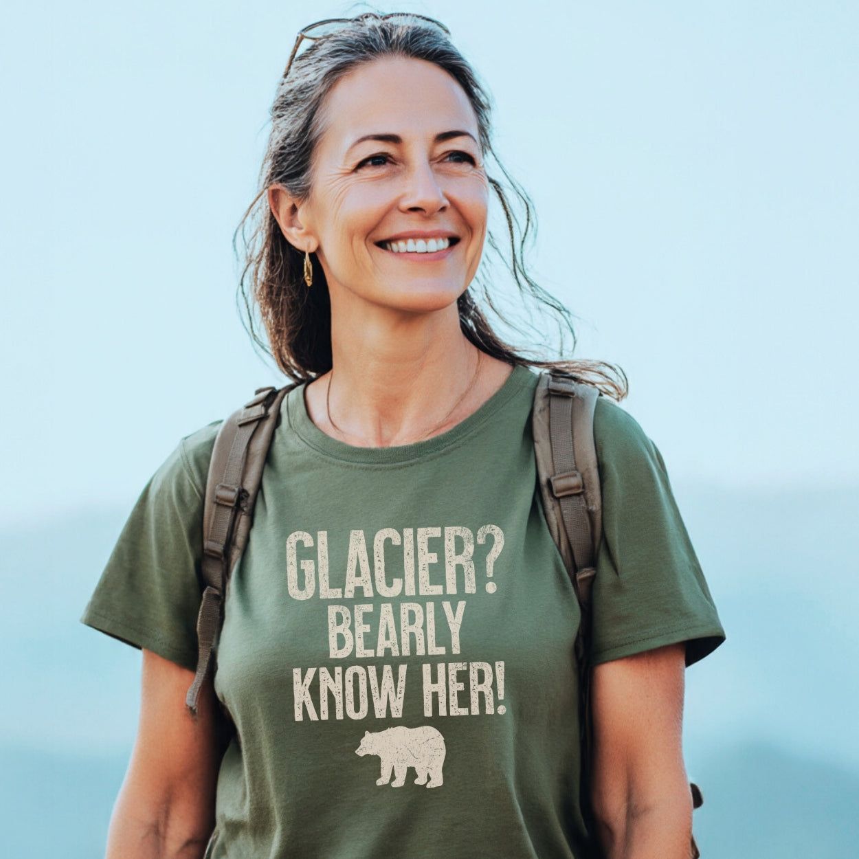 Middle-aged woman outdoors in a Glacier National Park Funny T-Shirt with a pun and bear silhouette, wearing a daypack, sunglasses, and earrings, embodying outdoor adventure and comfort.