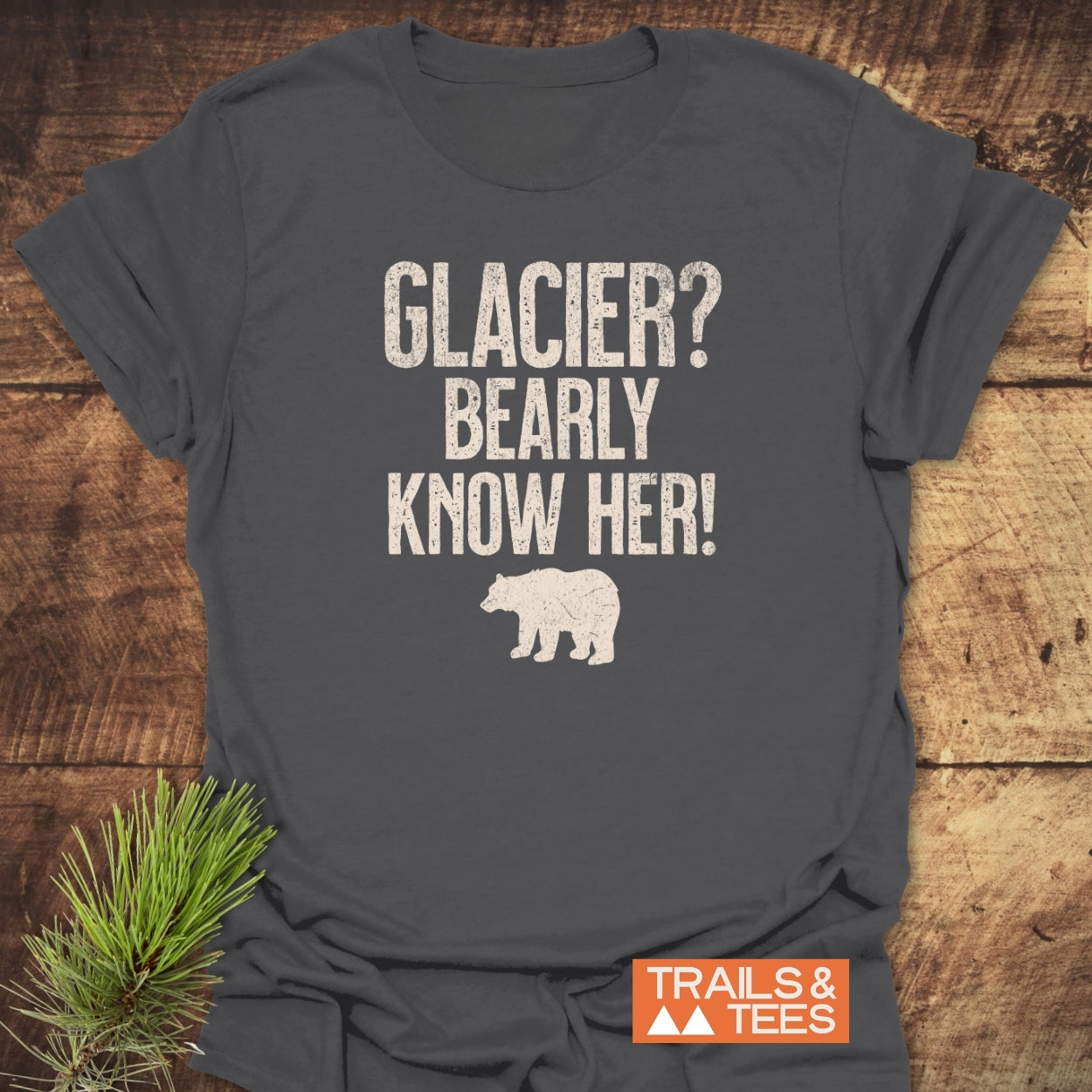 Charcoal-gray Glacier National Park Funny T-Shirt with GLACIER? BEARLY KNOW HER! and bear silhouette, laid on wood surface with pine sprig. Features durable print, soft cotton, and Trails & Tees logo.