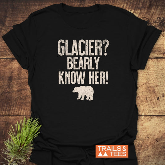 Glacier National Park Funny T-Shirt features a distressed white print with GLACIER? BEARLY KNOW HER! and a bear silhouette on a black tee, laid on wood with a pine sprig.