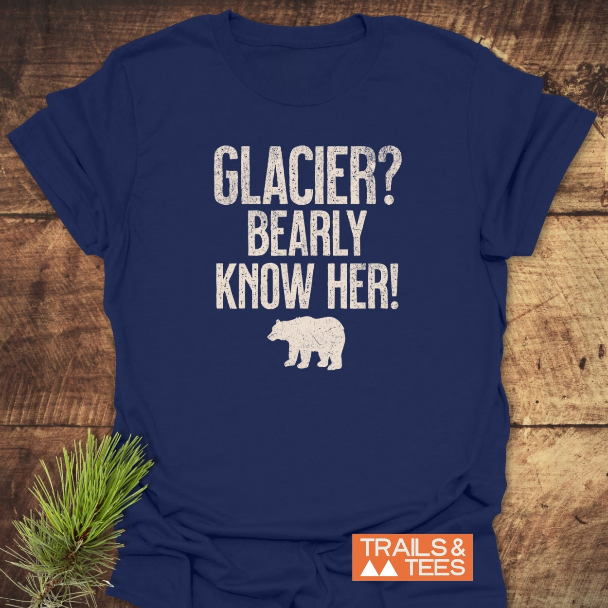 Navy-blue Glacier National Park Funny T-Shirt with distressed white print reading GLACIER? BEARLY KNOW HER! and a bear silhouette, laid on wood. Trails & Tees logo, pine greenery visible.