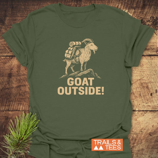 Goat Outside! Funny T-Shirt featuring a mountain goat with a hiking pack and slogan on an olive-green fabric; laid on wood, ideal for outdoor enthusiasts.