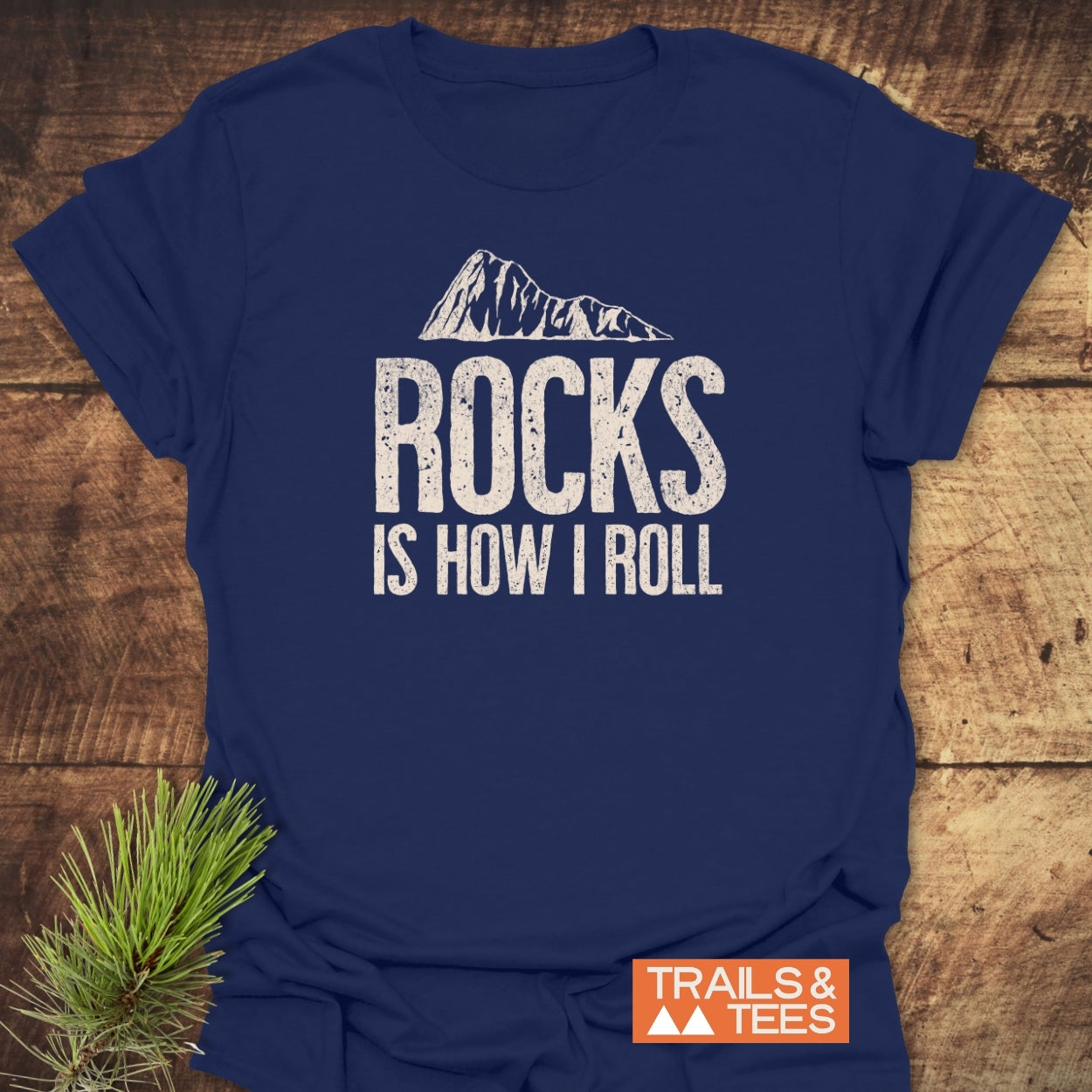 Navy-blue Rocks Is How I Roll T-shirt with mountain graphic, styled with pine bough, showcasing durable print and adventure-ready comfort from Trails & Tees.
