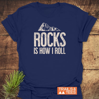 Navy-blue Rocks Is How I Roll T-shirt with mountain graphic, styled with pine bough, showcasing durable print and adventure-ready comfort from Trails & Tees.
