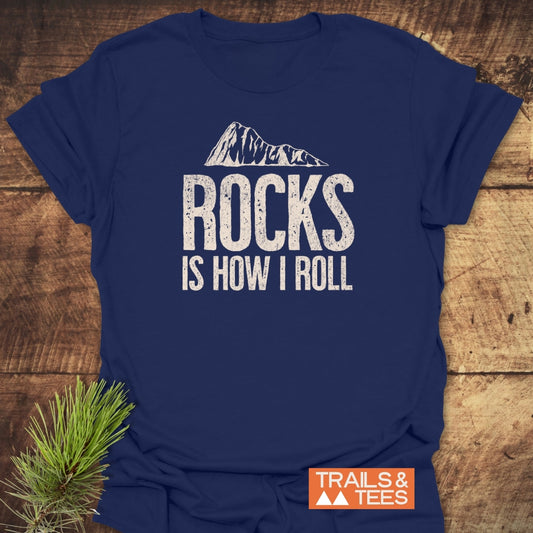 Navy-blue Rocks Is How I Roll T-shirt with mountain graphic, styled with pine bough, showcasing durable print and adventure-ready comfort from Trails & Tees.