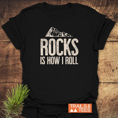 Rocks Is How I Roll T-Shirt featuring a mountain-outline graphic above the phrase, displayed on a wooden surface with a pine sprig, highlighting its outdoor-themed design and Trails & Tees logo.