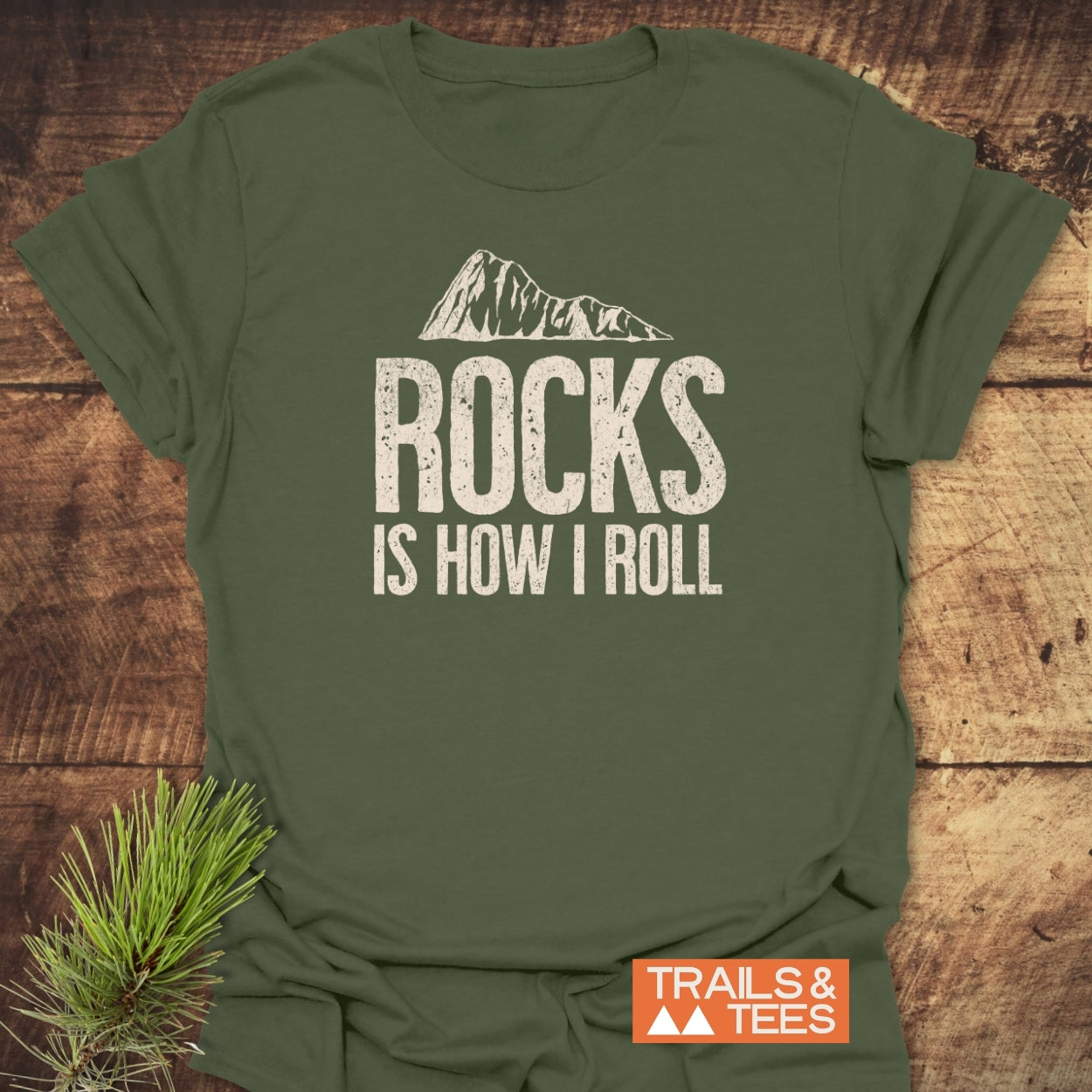 Forest-green Rocks Is How I Roll T-shirt with white mountain graphic, laid on rustic wood. Features TRAILS & TEES logo; pine sprig accents the outdoors theme.