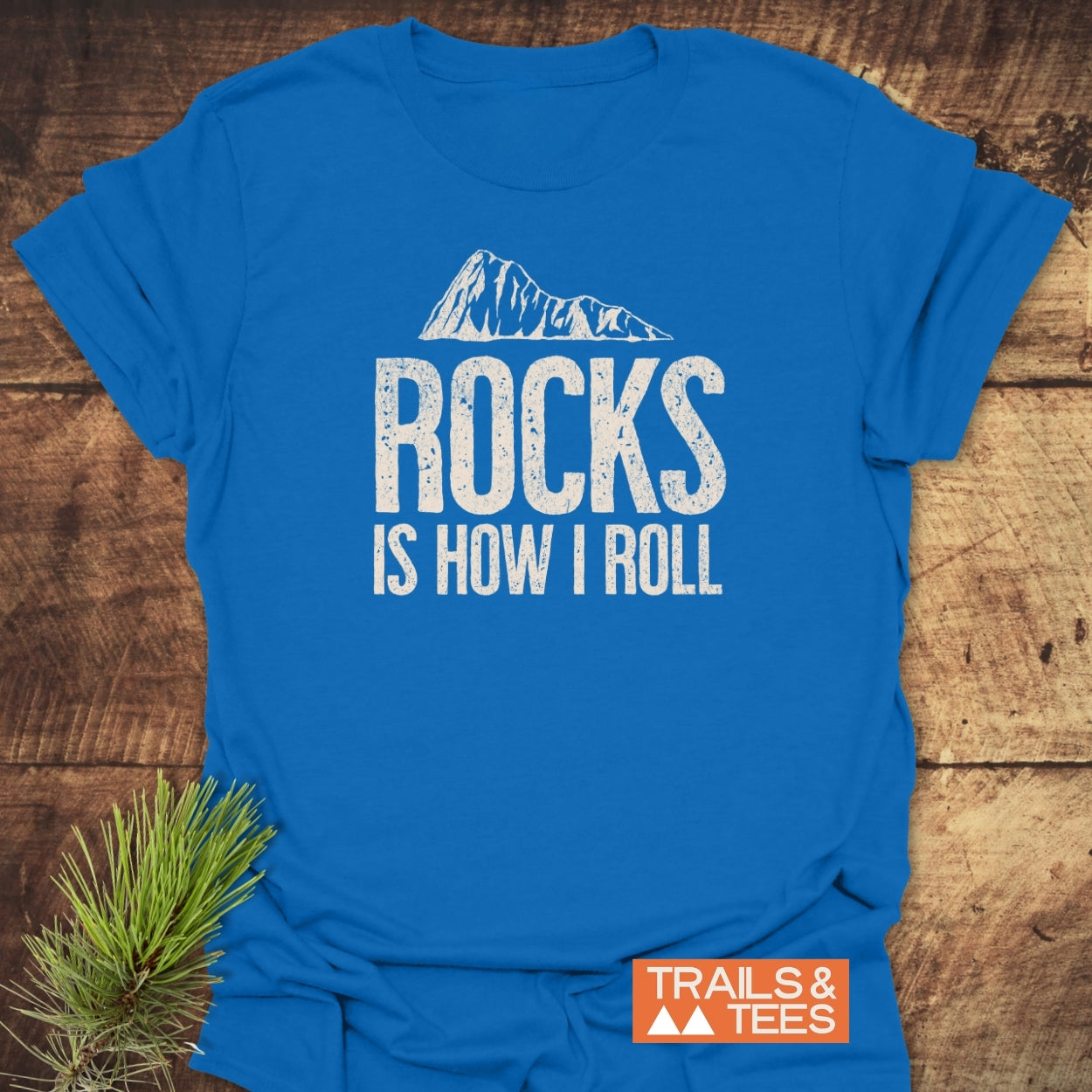 Rocks Is How I Roll T-Shirt with a stylized mountain graphic and bold lettering. Laid on wood with a pine needle accent, reflecting an outdoorsy vibe.