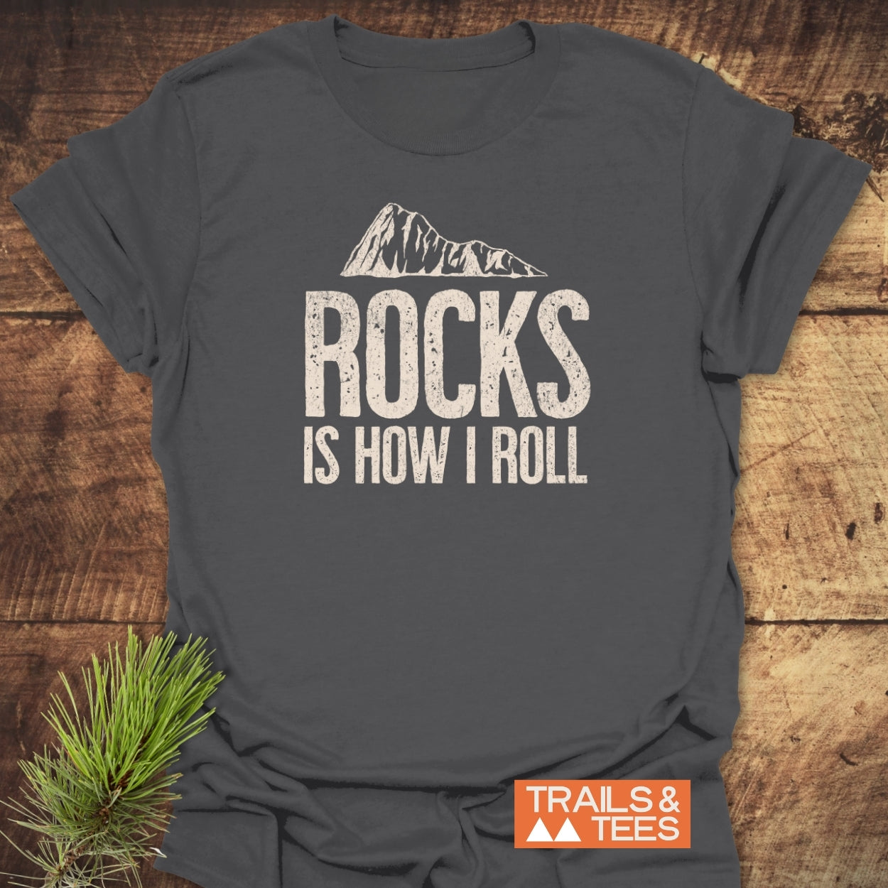 Rocks Is How I Roll T-Shirt with mountain peak graphic, emphasizing outdoor adventure. Laid on wood, features Trail & Tees logo. Pre-shrunk, ring-spun cotton for comfort and durability.
