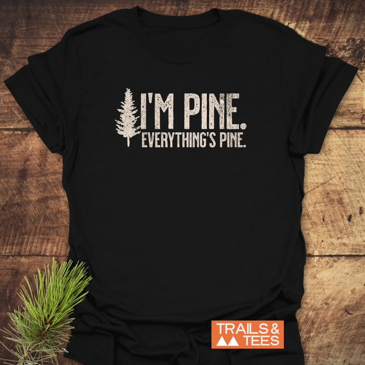 The I'm Pine. Everything's Pine. T-Shirt features a distressed white pine tree graphic and text on black fabric, enhanced by a decorative pine branch and TRAILS & TEES logo.
