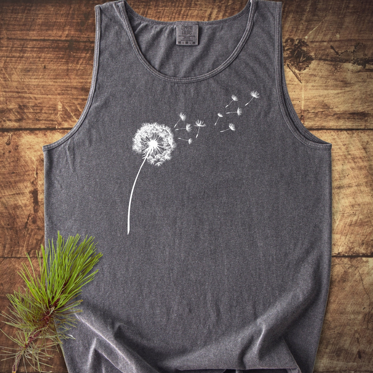 Dandelion Comfort Colors 9360 Tank Top, charcoal-gray, with a white dandelion design. Laid flat on wood, showcasing its relaxed fit and sustainable US cotton construction.