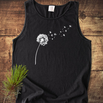 Dandelion Comfort Colors 9360 Tank Top features a black, sleeveless design with a white dandelion print. Laid on wood, it includes a pine sprig, highlighting its natural, sustainable appeal.