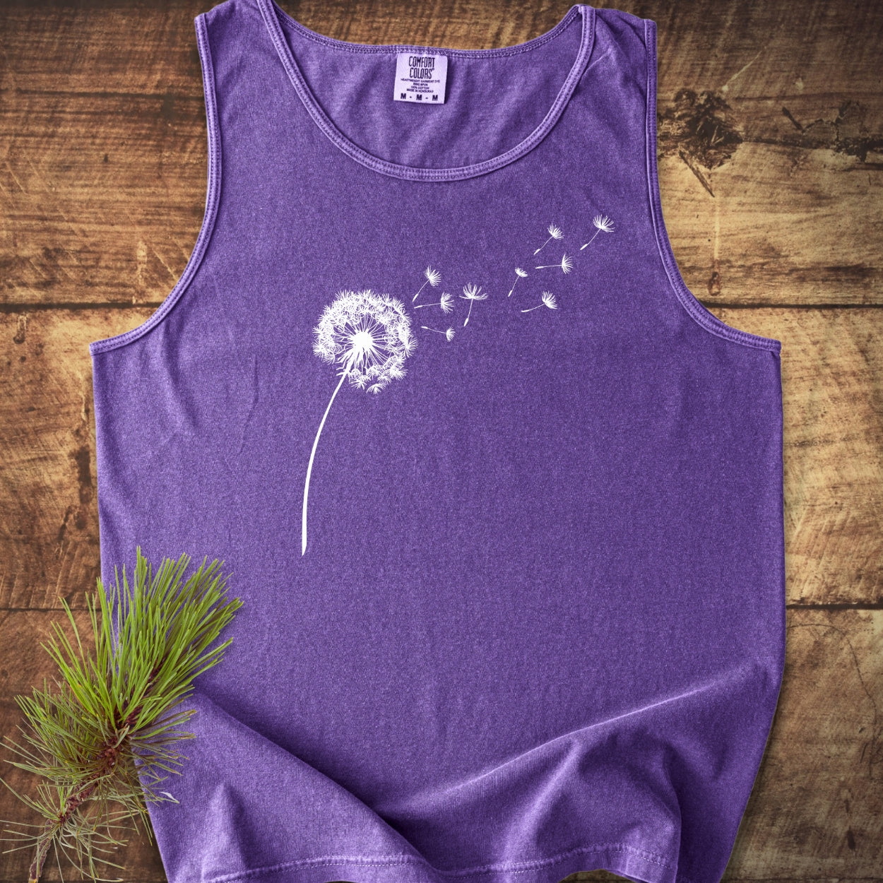 Dandelion Comfort Colors 9360 Tank Top featuring a white dandelion design, laid flat on wood with a pine branch accent; unisex, garment-dyed, relaxed fit, and made from sustainable US cotton.