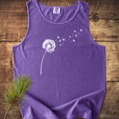 Dandelion Comfort Colors 9360 Tank Top featuring a white dandelion design, laid flat on wood with a pine branch accent; unisex, garment-dyed, relaxed fit, and made from sustainable US cotton.
