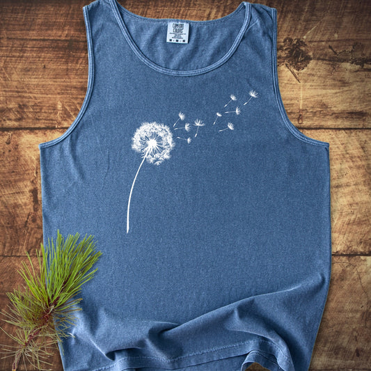Dandelion Comfort Colors 9360 Tank Top, sleeveless with a white dandelion graphic, laid flat on wood, showcasing its relaxed fit and eco-friendly craftsmanship, accented by a pine twig.