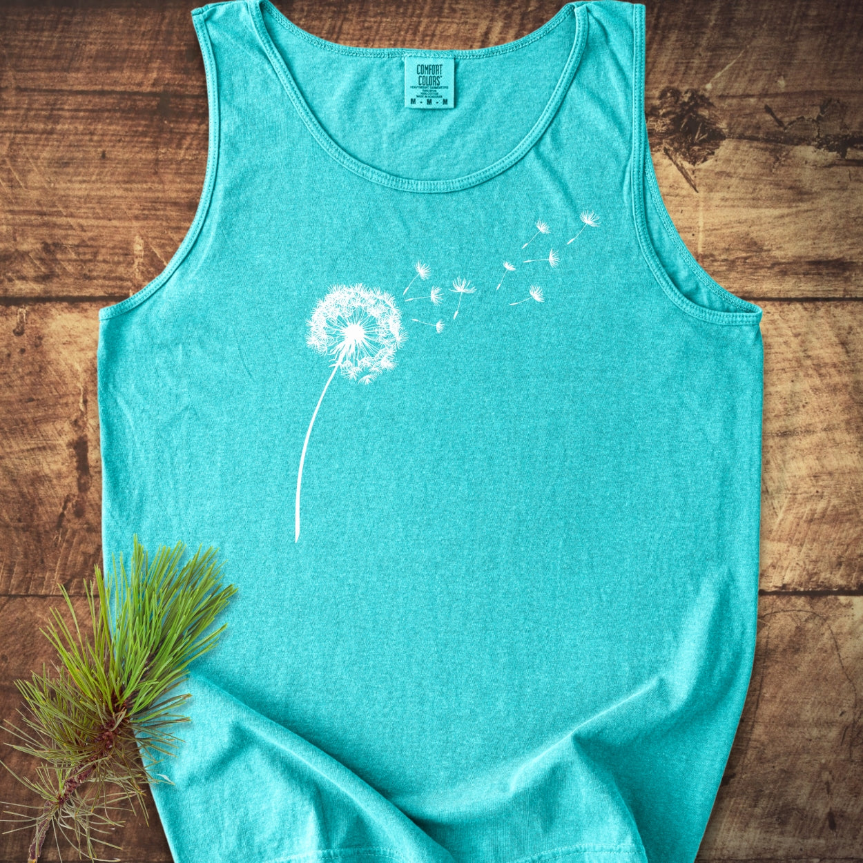 Dandelion Comfort Colors 9360 Tank Top features a white dandelion graphic, laid flat on wood with a pine branch beside it, highlighting its relaxed fit and sustainable cotton fabric.