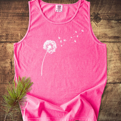 Dandelion Comfort Colors 9360 Tank Top, pink and sleeveless, features a white dandelion graphic with seeds drifting away. It's laid on wood, accompanied by a pine branch for a natural touch.